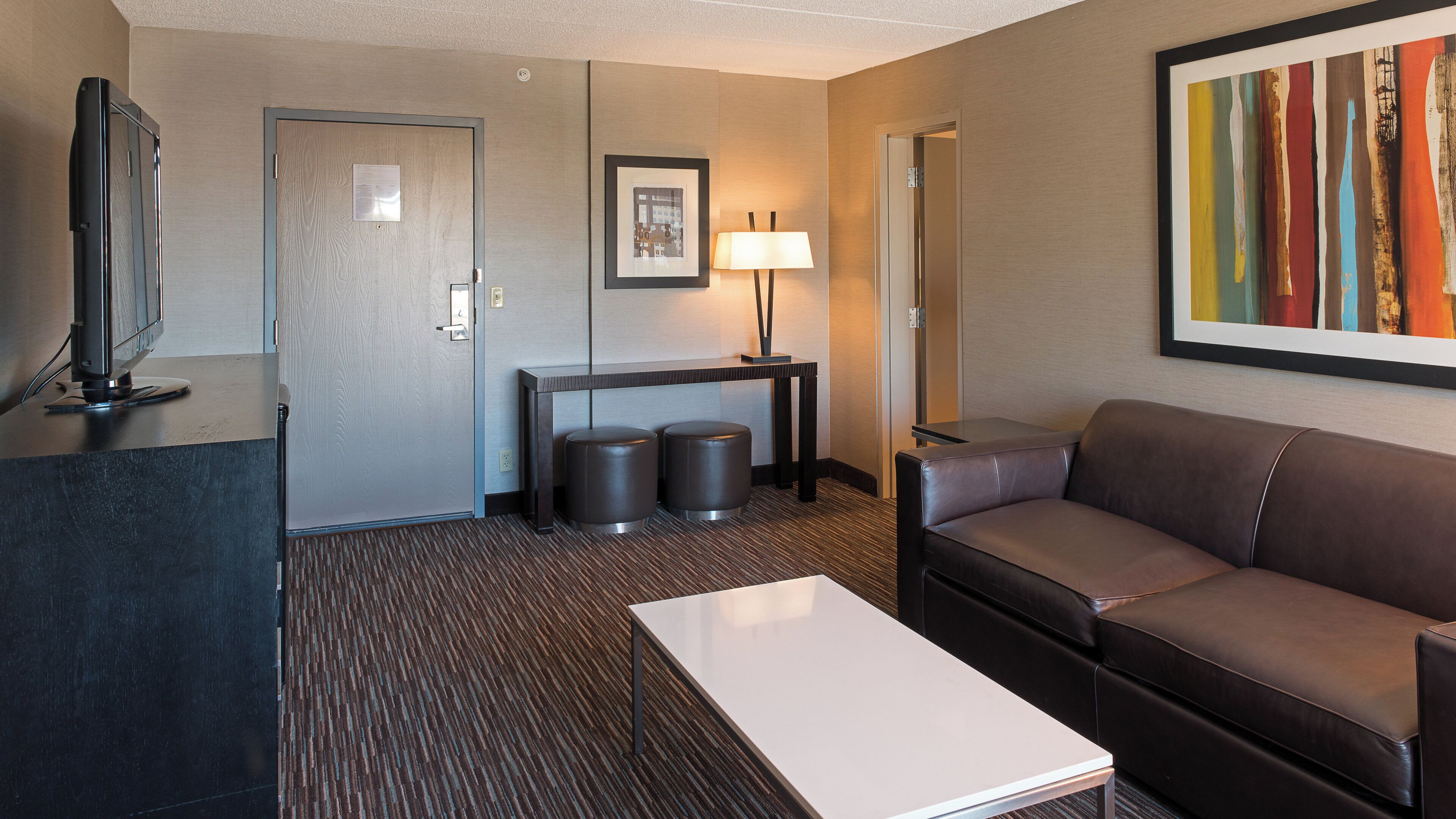 A hotel room with a flat-screen TV, a sofa, a coffee table, a desk with two stools, a lamp, and a framed picture on the wall.