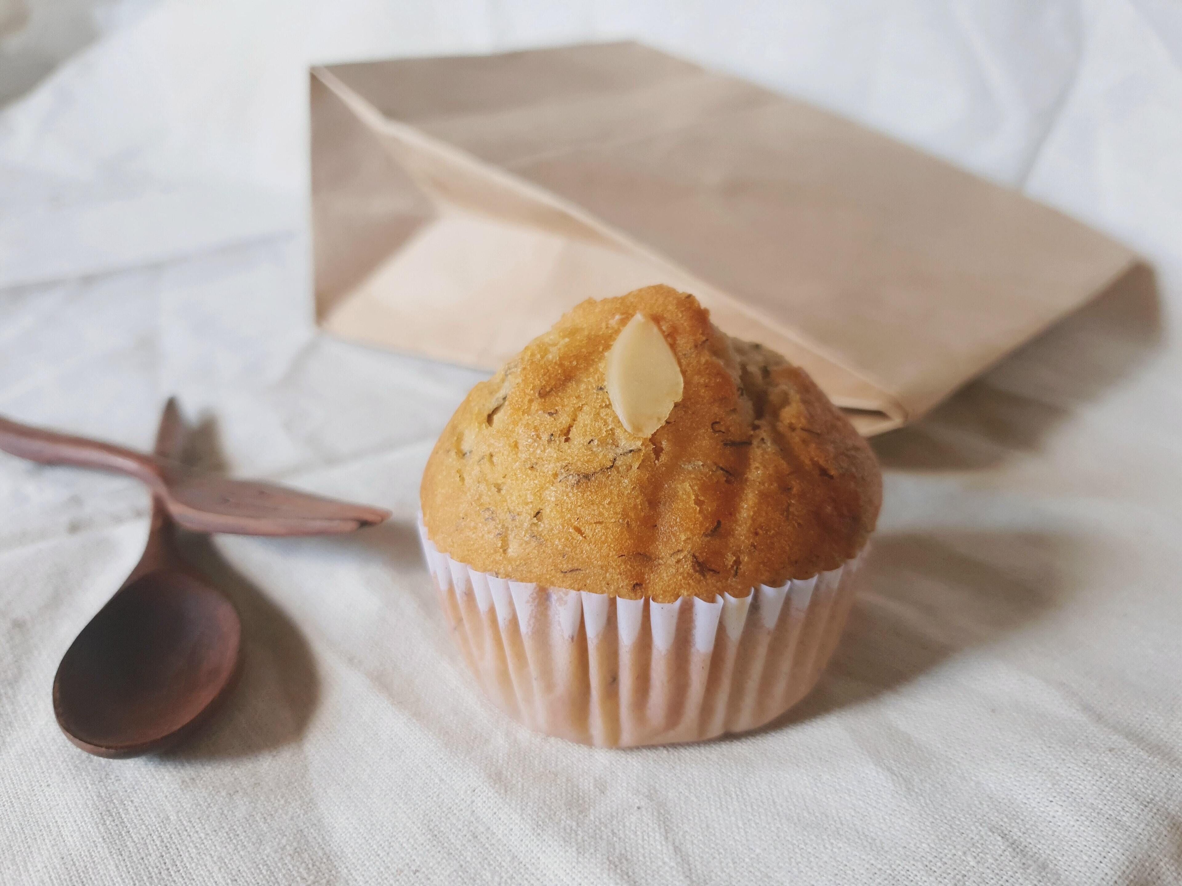 A muffin in a paper cup on a white cloth with a brown paper bag and a spoon in the background.