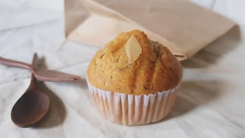 Un muffin en un got de paper damunt d'un drap blanc, amb una bossa de paper marró i una cullera al fons.