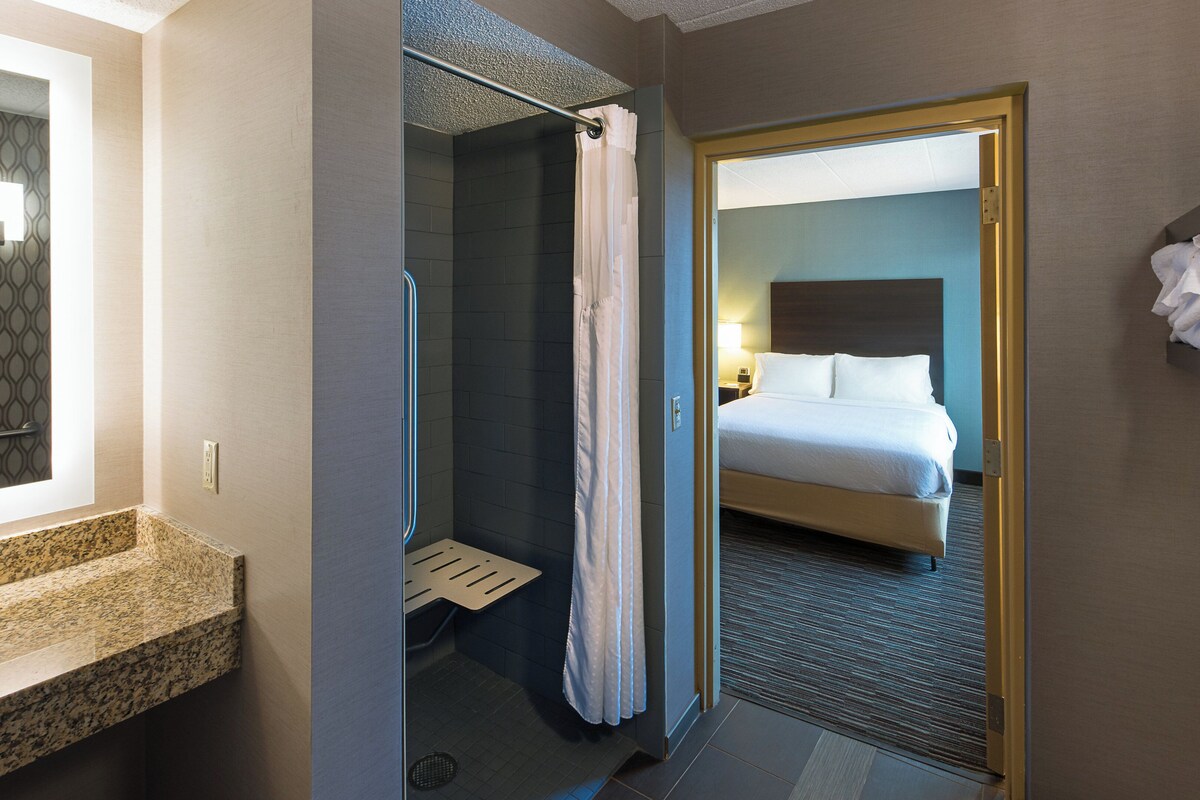 Habitación de hotel con ducha, cama, baño con lavamanos y toallero.