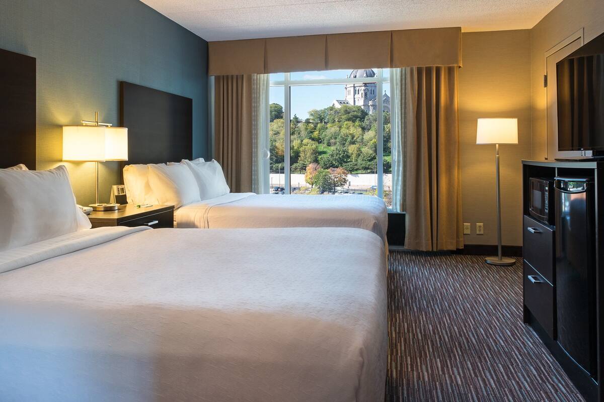 Ein Hotelzimmer mit zwei Betten, einem Schreibtisch, einer Lampe, einem Fernseher und Blick auf ein Gebäude.