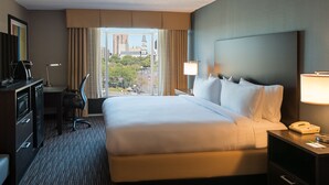 A hotel room with a large bed, a desk with a lamp, a telephone, and a view of the cityscape through the window.