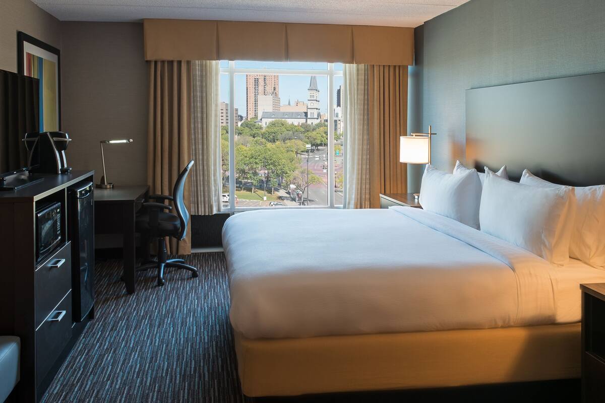 Ein Hotelzimmer mit einem großen Bett, einem Schreibtisch mit Lampe, einem Telefon und Blick auf die Stadt durch das Fenster.