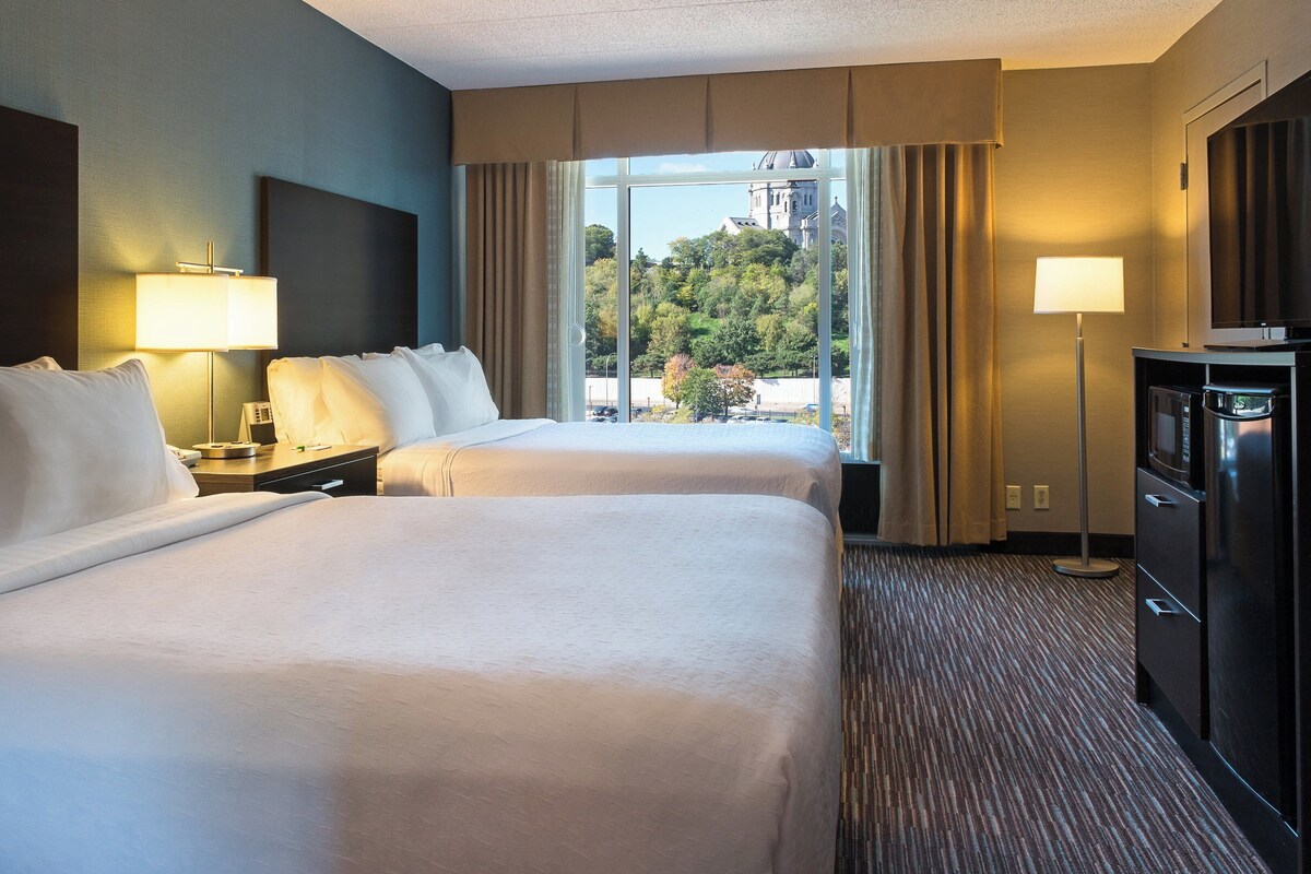 Habitación de hotel con dos camas, un escritorio con lámpara, televisión y vista a un edificio a través del ventanal.