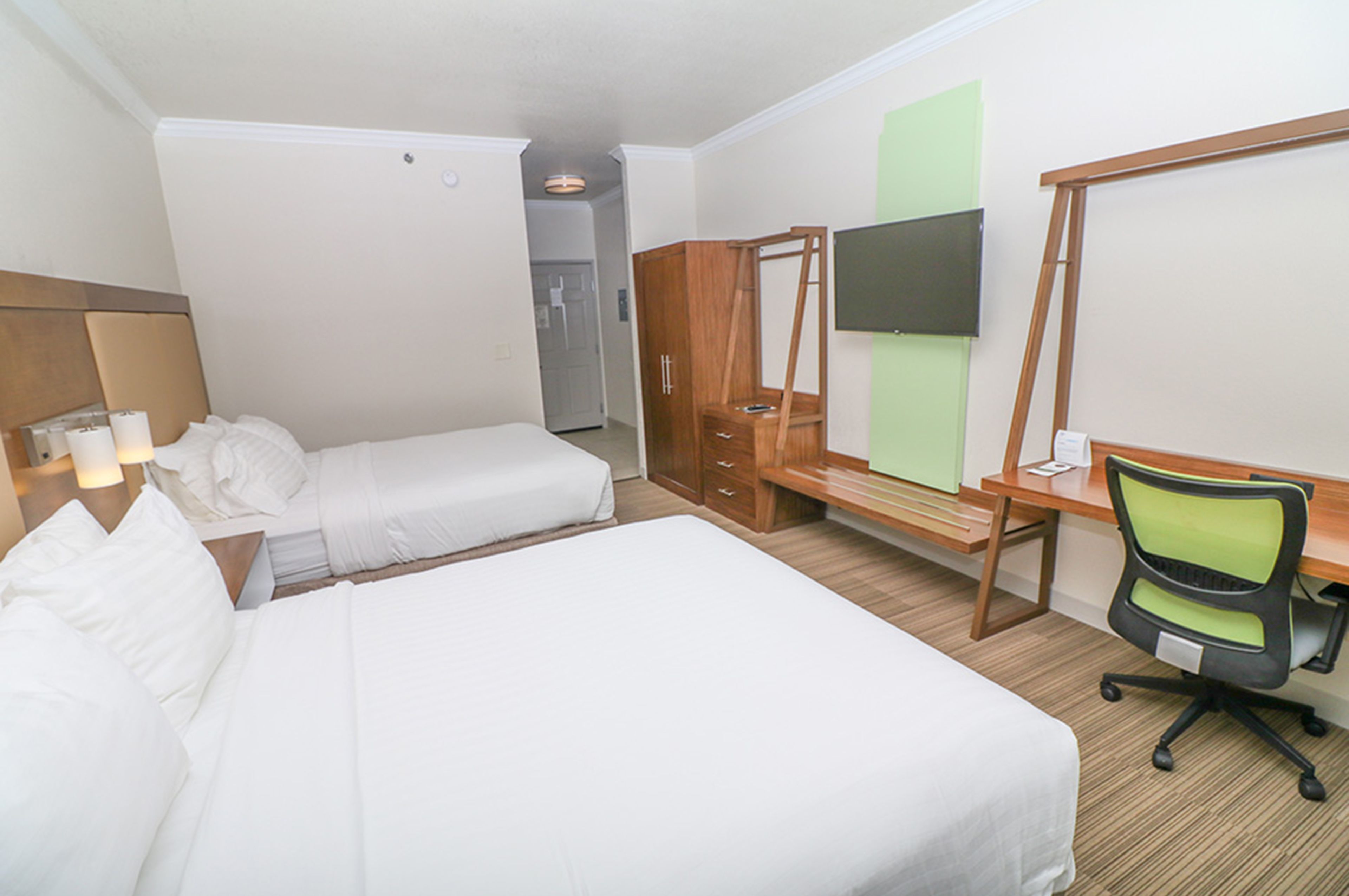 A hotel room with a large bed, a desk with a chair, a wardrobe, and a flat-screen TV mounted on the wall.