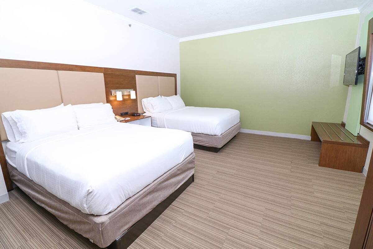 A hotel room with two beds, a wooden headboard, a small wooden bench, a nightstand, and a wall-mounted TV.