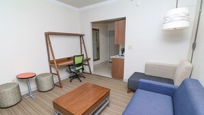A compact living space with a sofa, a small table, a desk with a chair, and a kitchenette area.