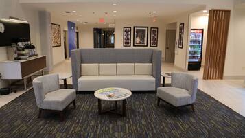 A lobby with a sofa, two armchairs, a coffee station, a vending machine, and framed artwork on the walls.