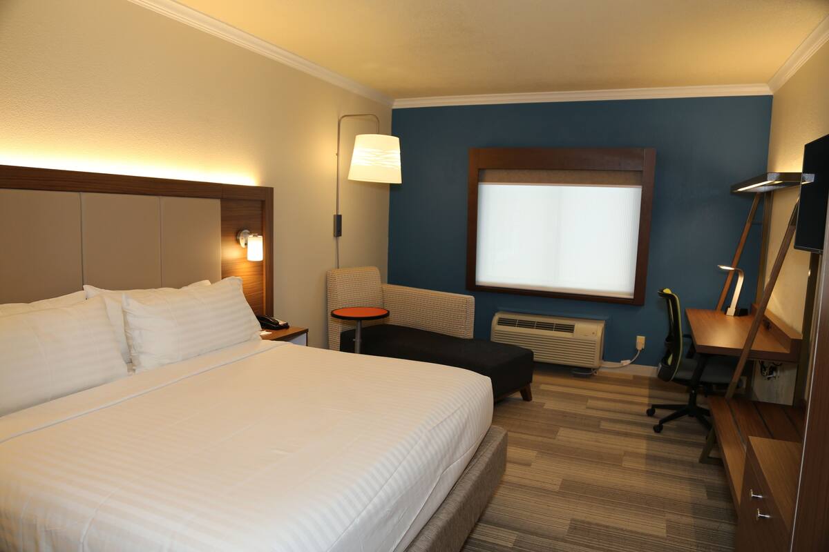 A hotel room with a large bed, a desk, a chair, a small table, a lamp, and a window.