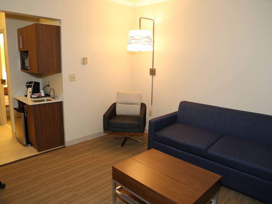 A modern living room with a blue sofa, a wooden coffee table, a patterned armchair, and a kitchenette area with a microwave and coffee machine.