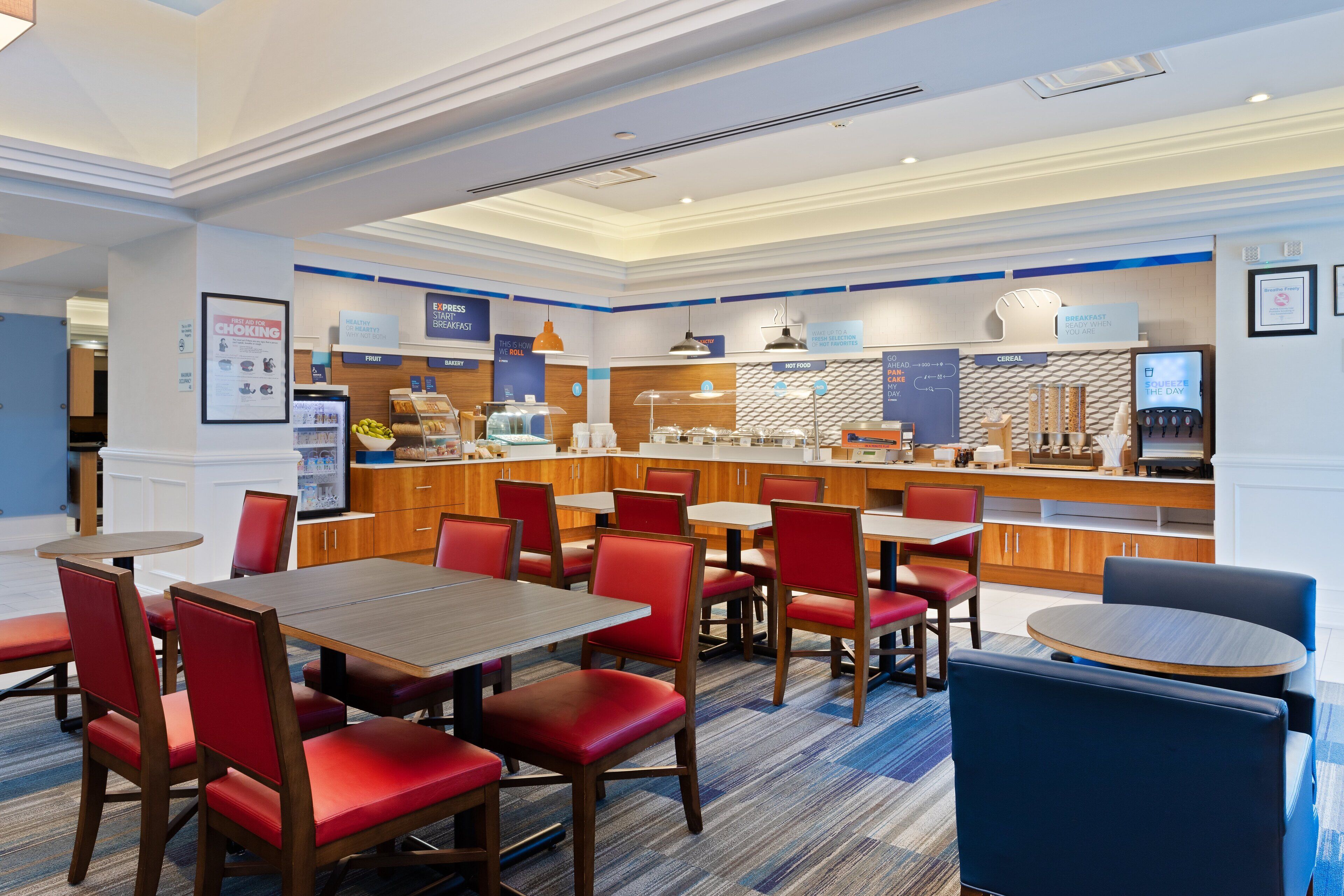 A hotel restaurant with red upholstered chairs, wooden tables, and a breakfast bar.