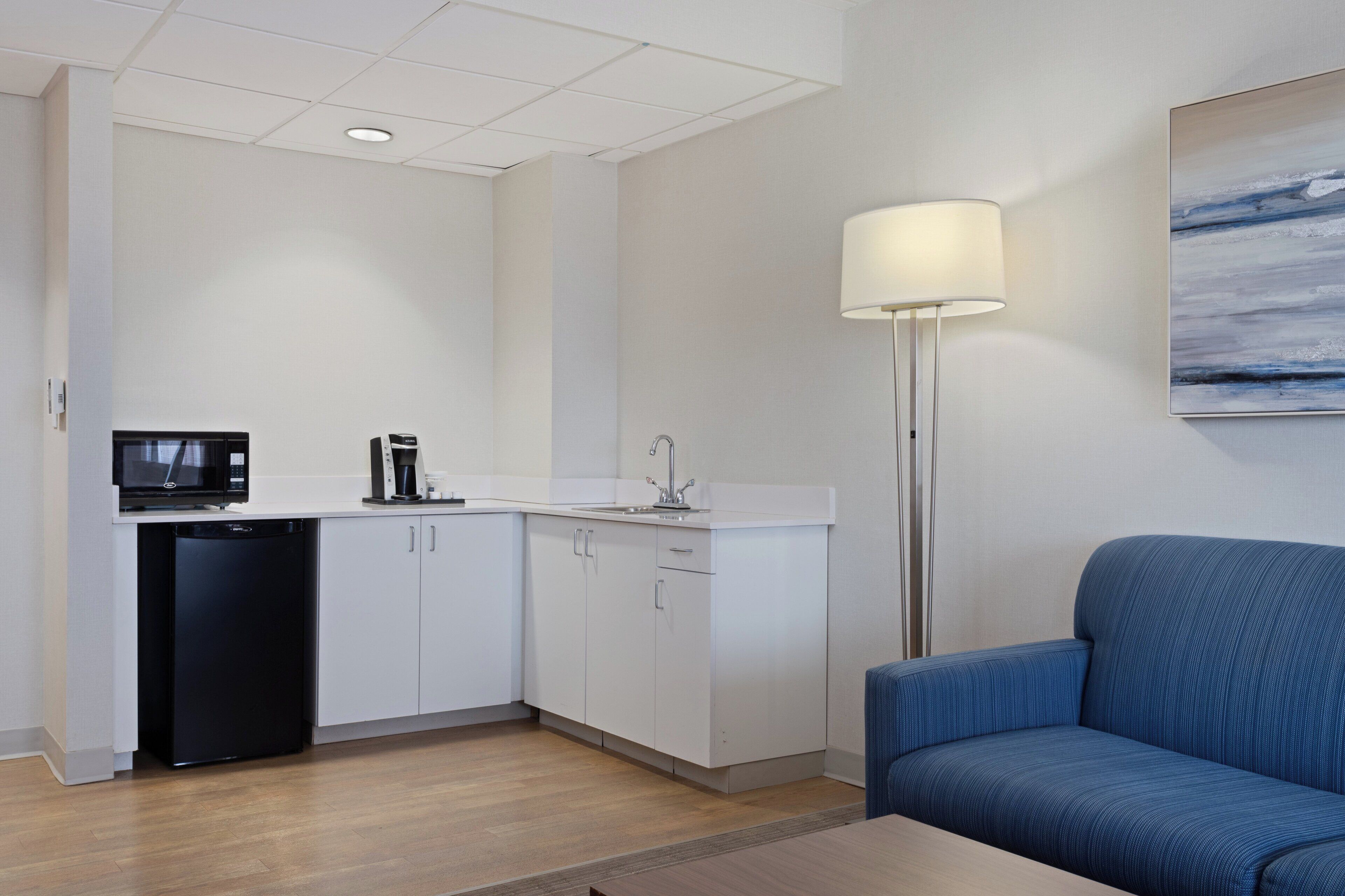 A modern kitchen with white cabinets, a black dishwasher, and a microwave. There is a blue sofa and a floor lamp in the living area.