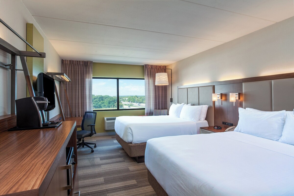 A hotel room with two beds, a desk with a computer, a chair, and a window with a view.