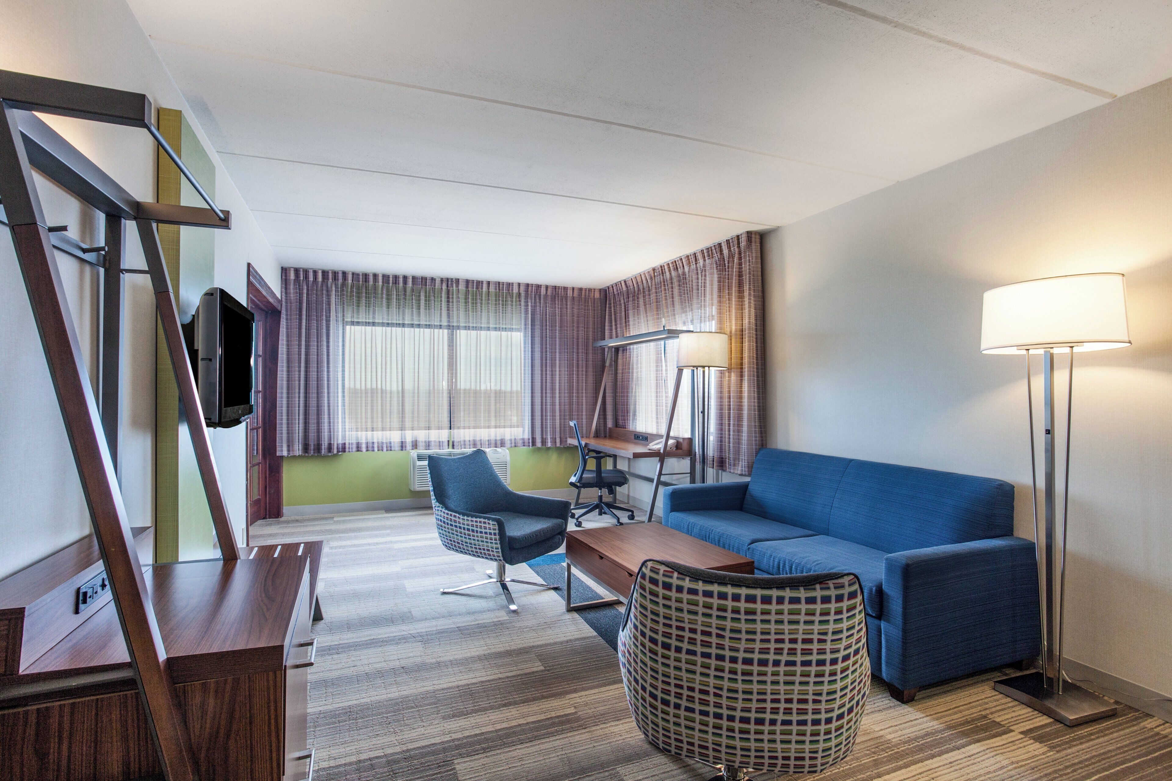 A hotel room with a sofa, a desk, a chair, a television, a lamp, and a patterned ottoman.