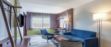 Habitación de hotel con sofá, escritorio, silla, televisión, lámpara y una otomana estampada.