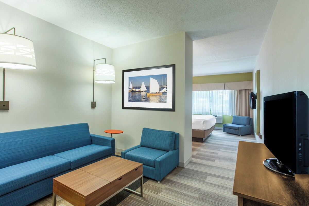 A hotel room with a blue sofa, a small blue armchair, a wooden coffee table, a framed picture of sailboats, a television, and a bed with a blue headboard.