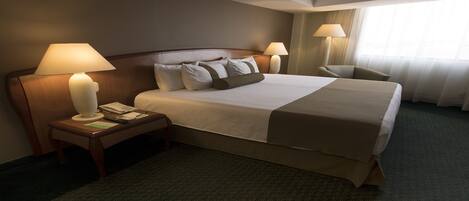 Habitación de hotel con una cama grande, dos mesitas de noche con lámparas, una silla y una ventana con cortinas.