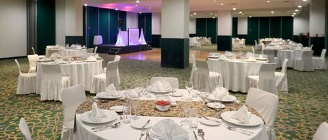 Un salón de banquetes con mesas redondas dispuestas para cenar, un escenario con iluminación púrpura y un suelo cubierto de alfombra verde.