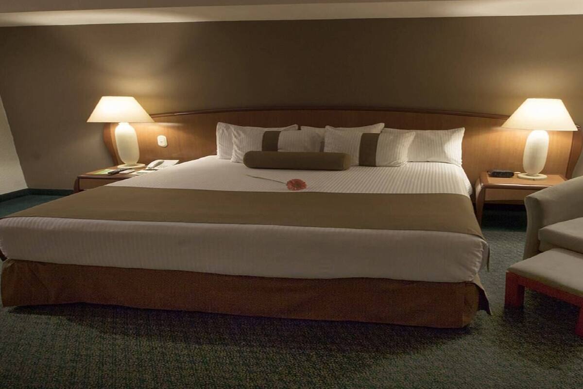Una habitación de hotel con una cama grande, dos lámparas de noche, una silla y una mesita.