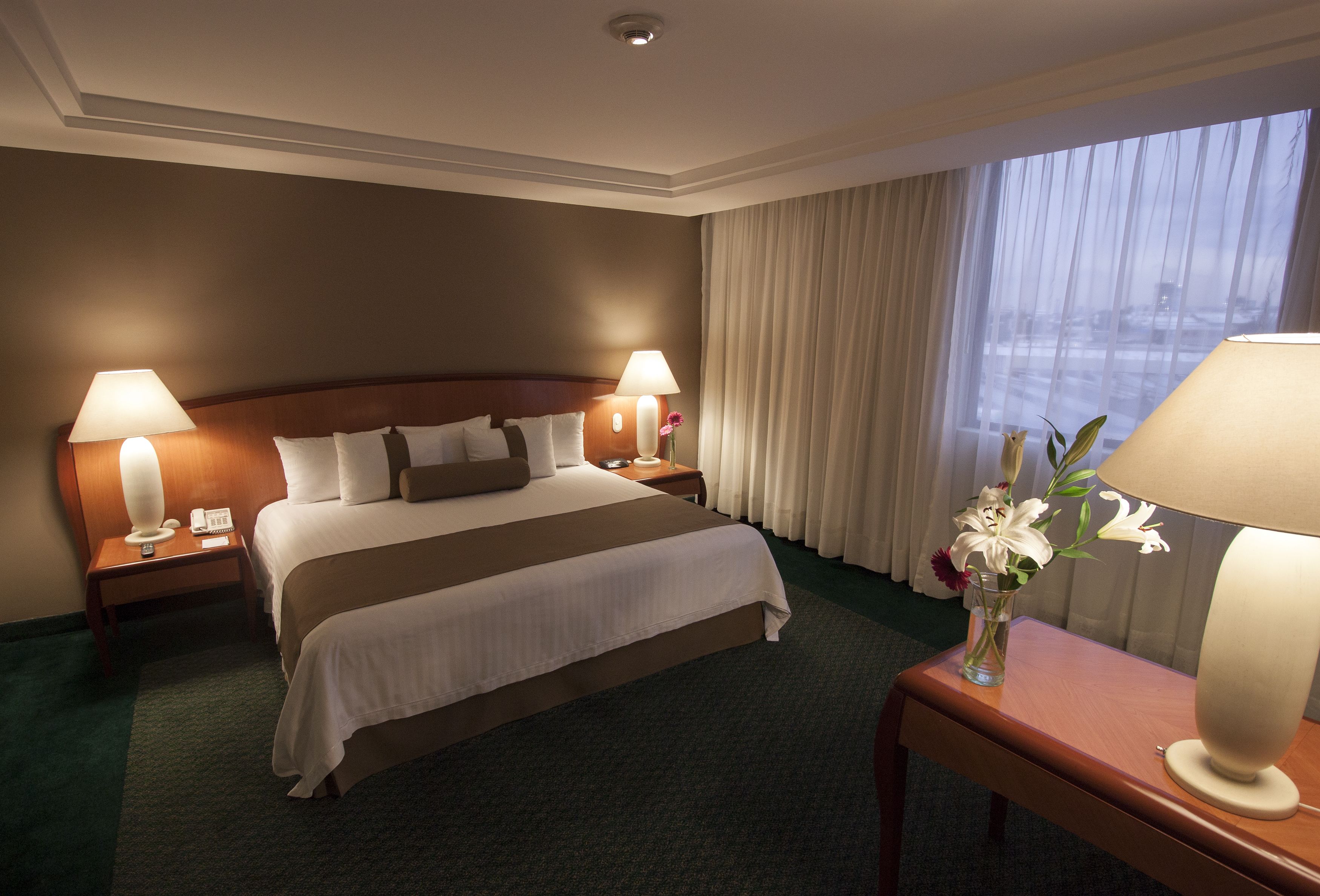 A hotel room with a large bed, two bedside lamps, a vase with flowers, and a view of the city through the window.