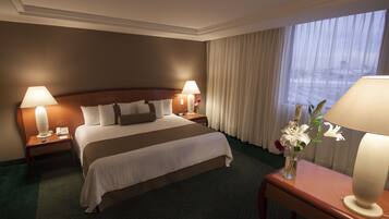 A hotel room with a large bed, two bedside lamps, a vase with flowers, and a view of the city through the window.