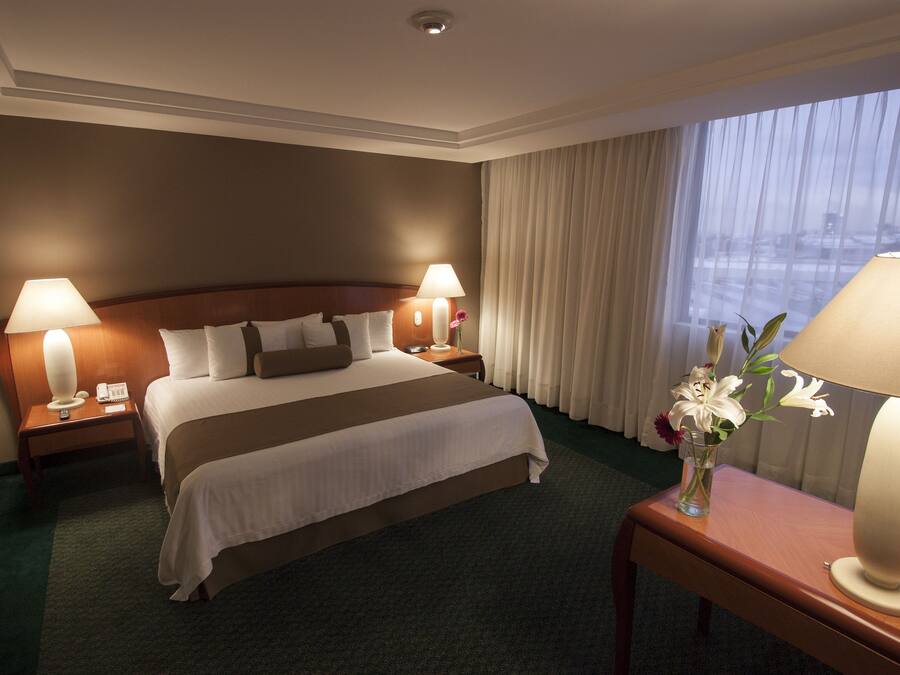 A hotel room with a large bed, two bedside lamps, a vase with flowers, and a view of the city through the window.