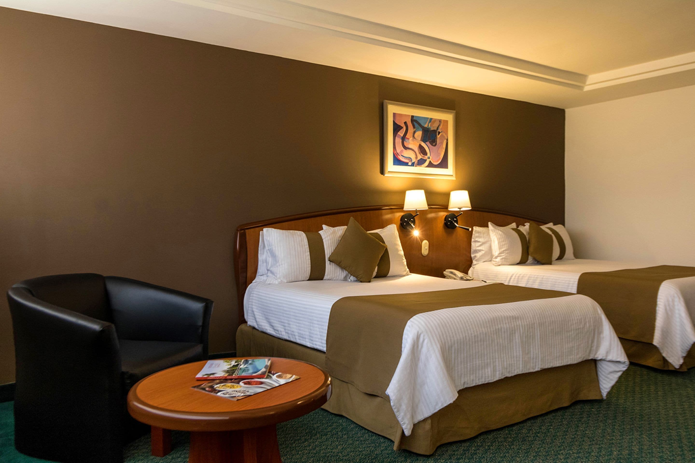 A hotel room with two beds, a chair, a table, and a painting on the wall.
