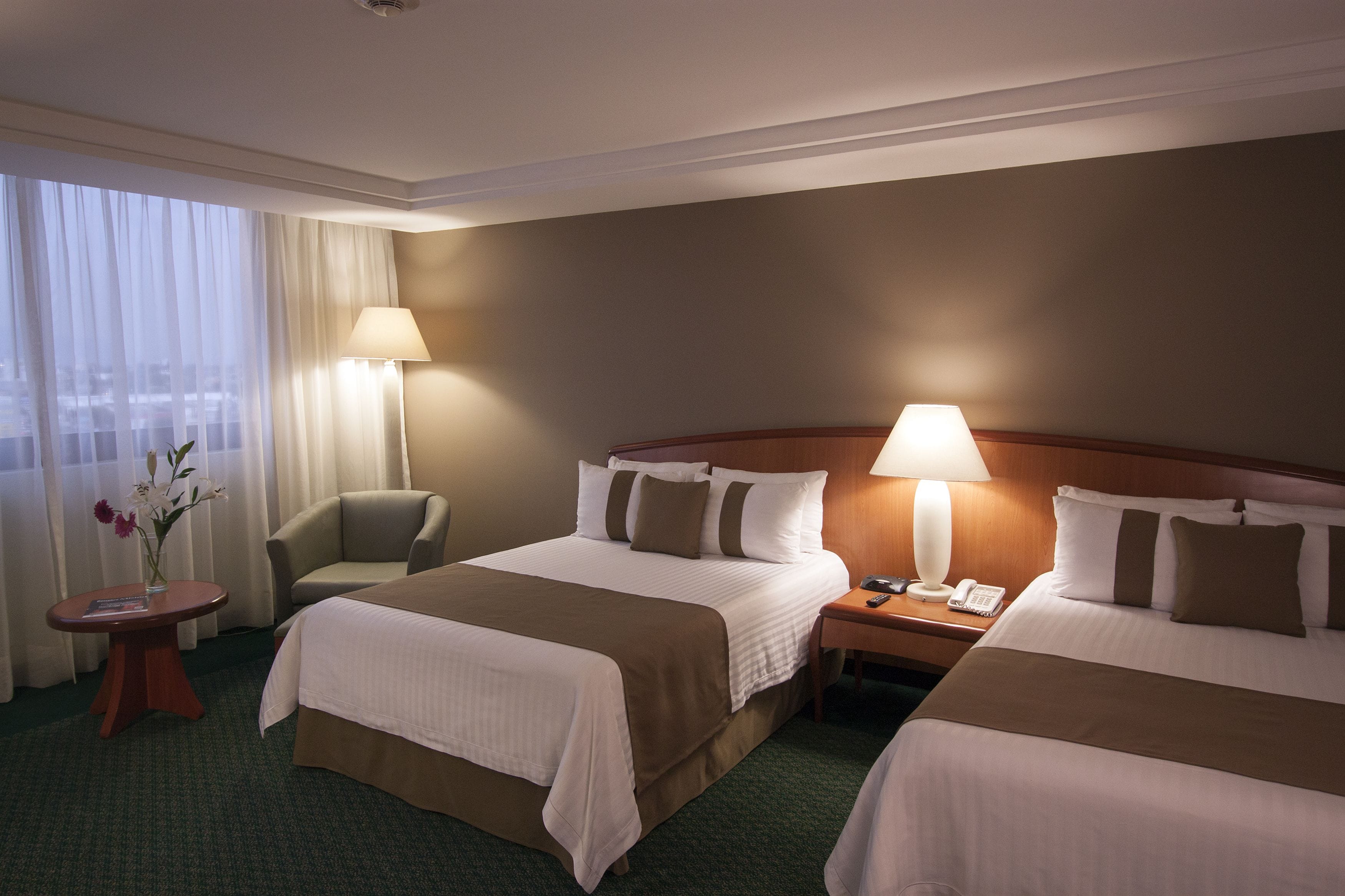 A hotel room with two beds, a chair, a table with a vase of flowers, and a lamp on each side of the bed.