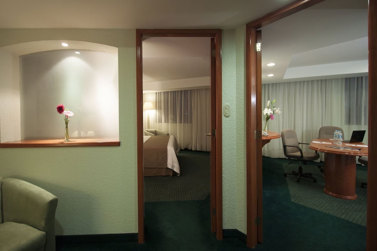 Une chambre d’hôtel avec un lit, un bureau avec un ordinateur portable, une chaise et une petite table sur laquelle est posé un vase de fleurs.