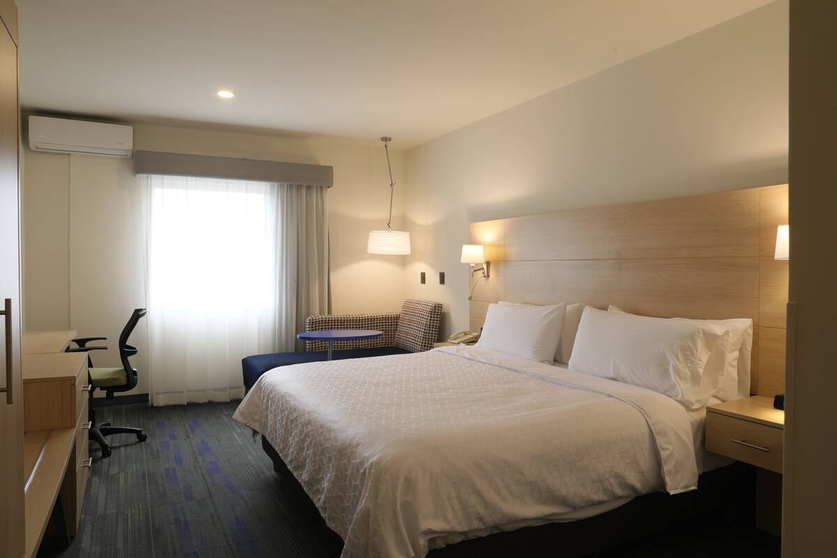 A hotel room with a large bed, a desk, a chair, a small table, and a window with curtains.