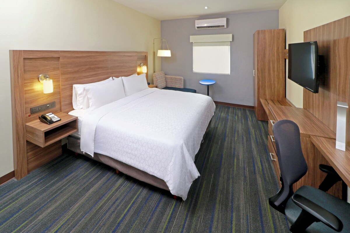 Habitación de hotel con una cama grande, un escritorio con silla, un televisor de pantalla plana, una mesita de noche con lámpara y una ventana.