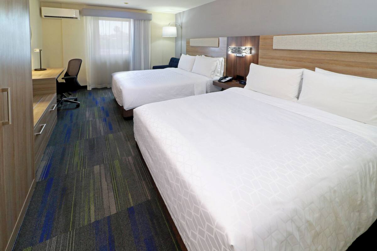 A hotel room with two beds, a desk, a chair, a lamp, and a window with curtains.