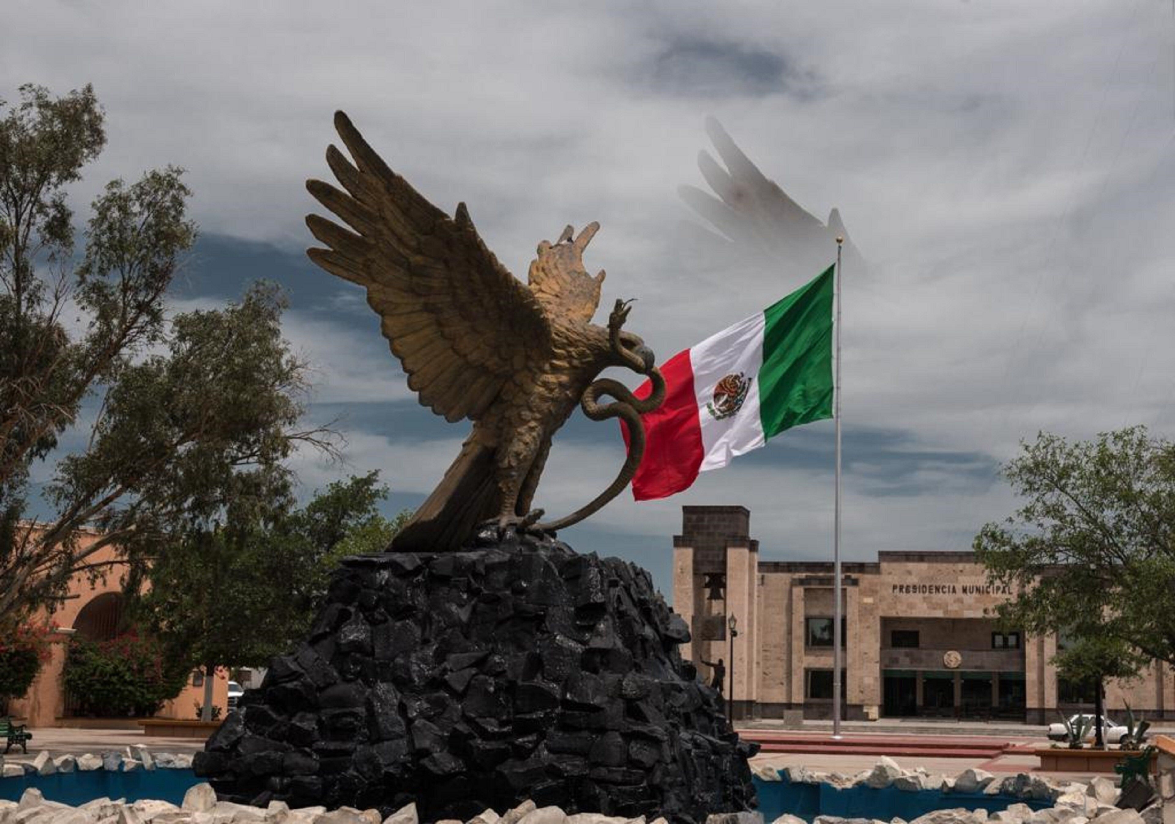 A statue of an eagle with a flag on its back, set on a rocky base, with a building in the background.