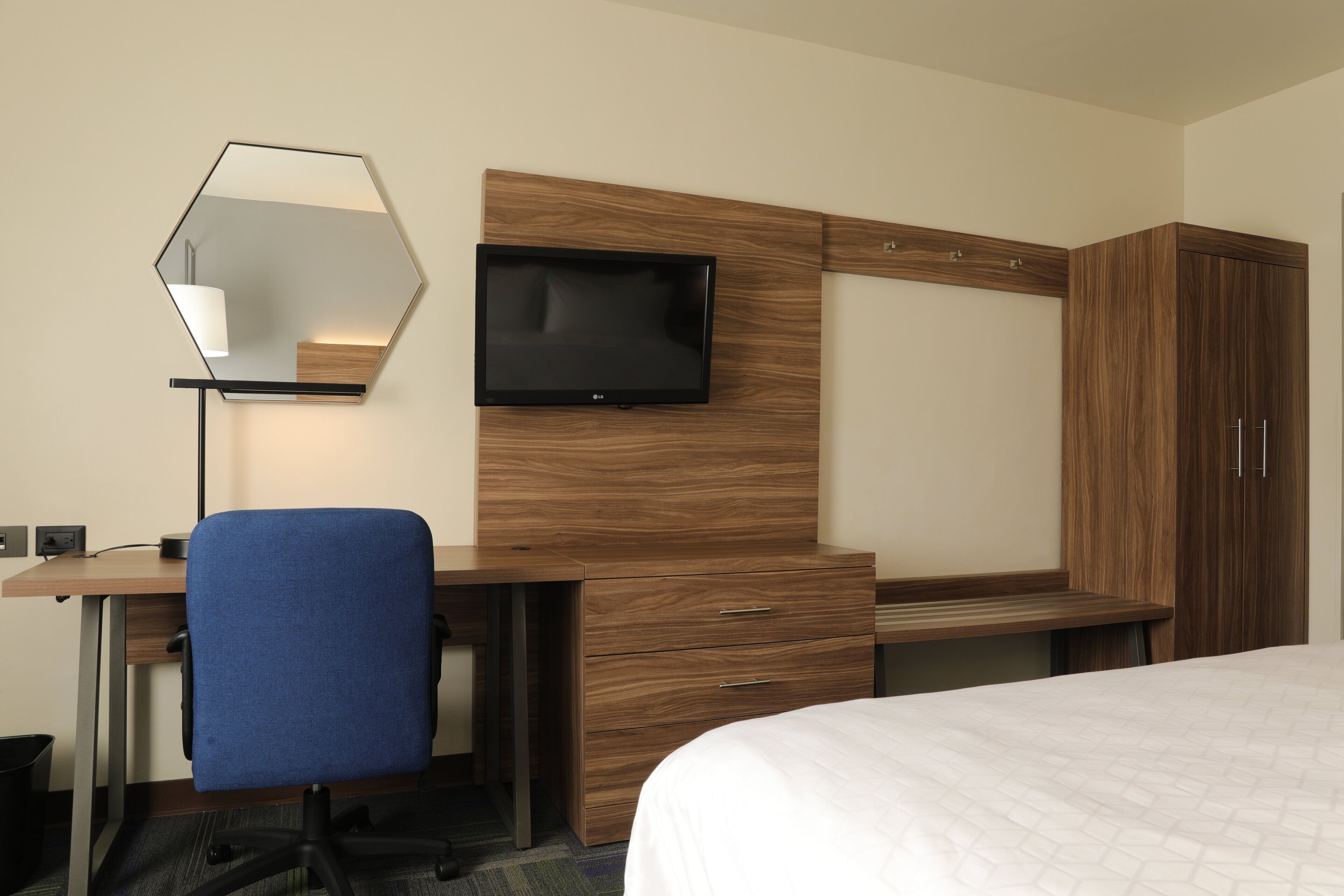 Habitación de hotel con un escritorio de madera, una silla de oficina azul, un televisor de pantalla plana, un armario y una cama con colcha blanca.