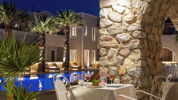 A poolside dining area with stone walls, palm trees, and a view of a building with a dome. - Rosebay Hotel (Santorini)
