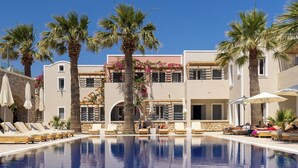 A swimming pool with lounge chairs and palm trees, surrounded by buildings with balconies and flower arrangements. - Rosebay Hotel (Santorini)