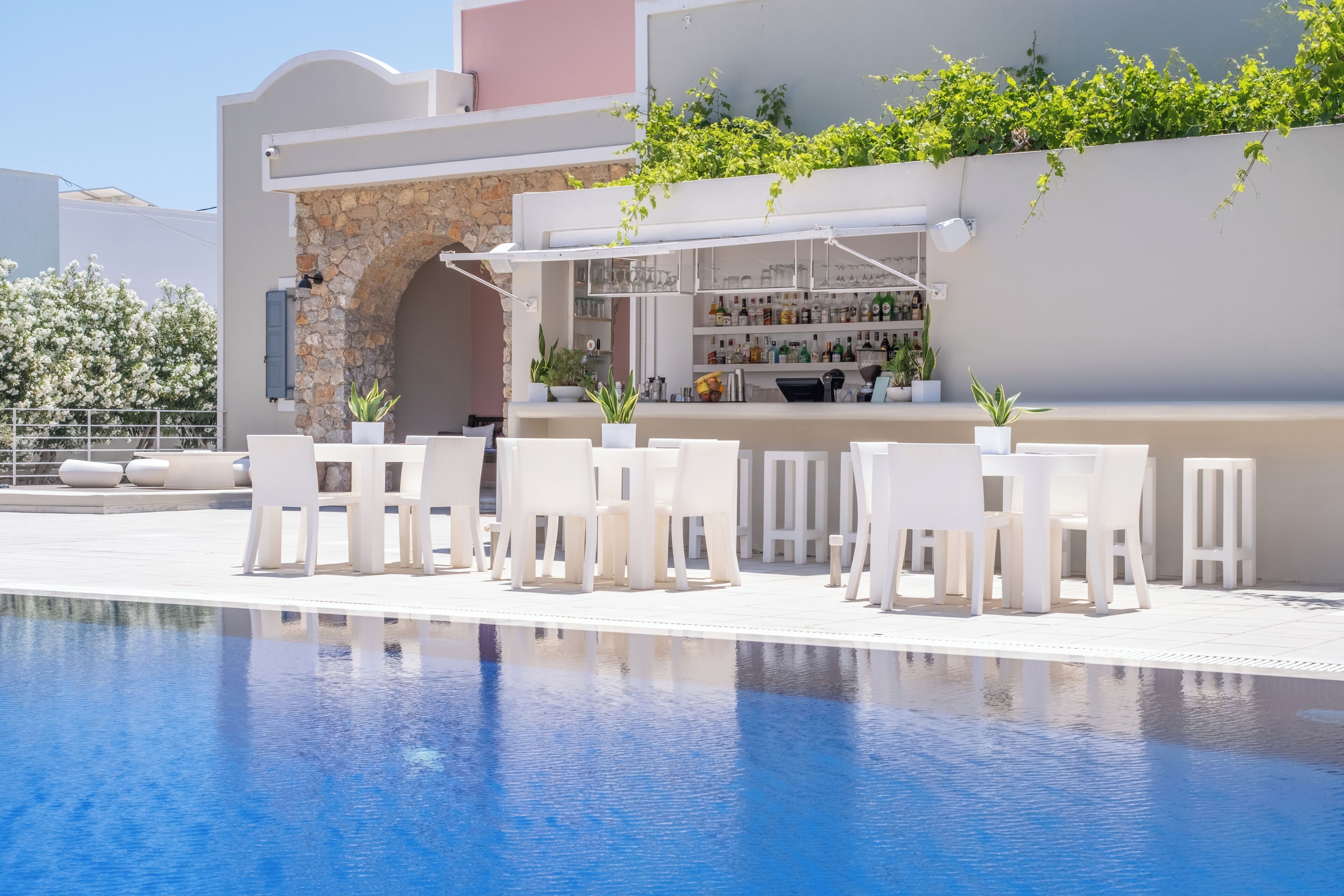 Un espace aménagé au bord de la piscine, avec des chaises et des tables blanches, un bar doté d’étagères remplies de bouteilles et un mur en pierre.