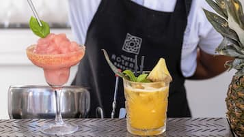 A bar setting with a drink in a margarita glass garnished with a leaf, a cocktail in a glass with a pineapple slice, and a pineapple on the counter.