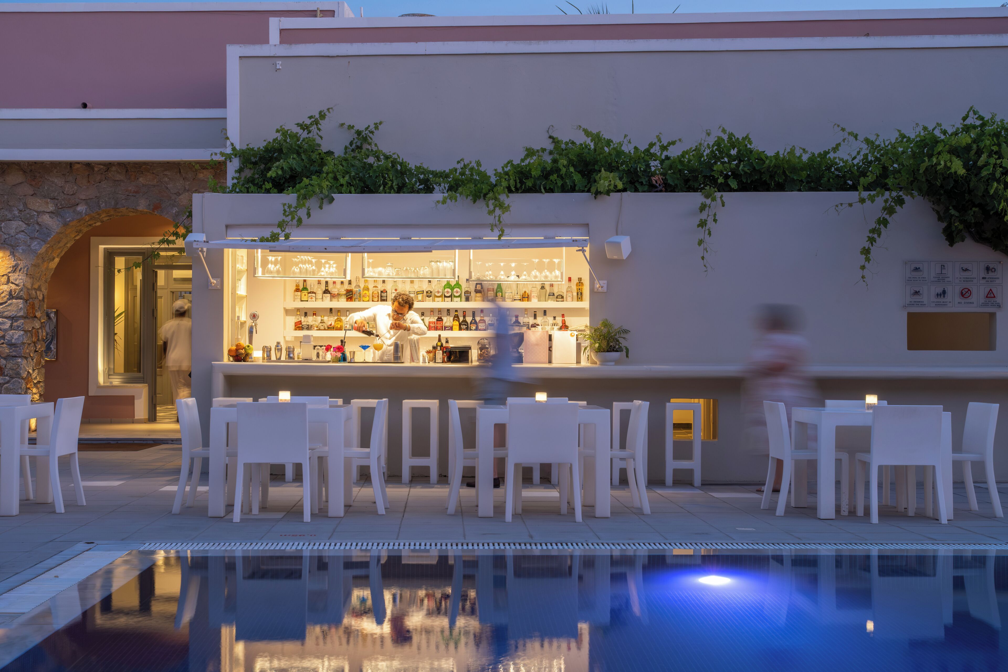 A poolside bar with white chairs and tables, a bartender, and a stone wall with greenery.