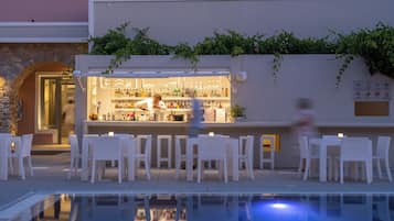 A poolside bar with white chairs and tables, a bartender, and a stone wall with greenery.