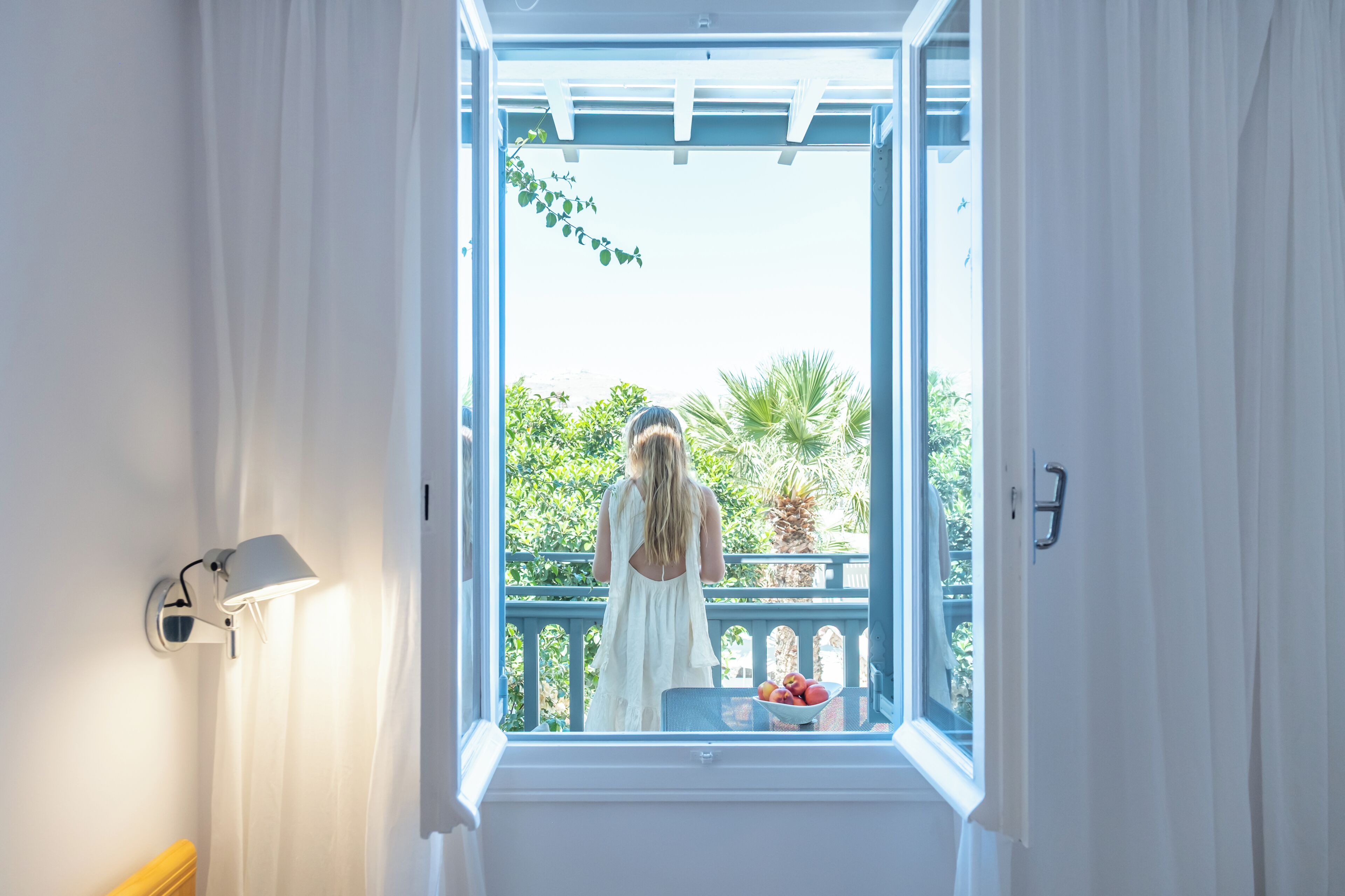 Et værelse med et vindue til en balkon med udsigt over grøn vegetation og en skål med frugt.