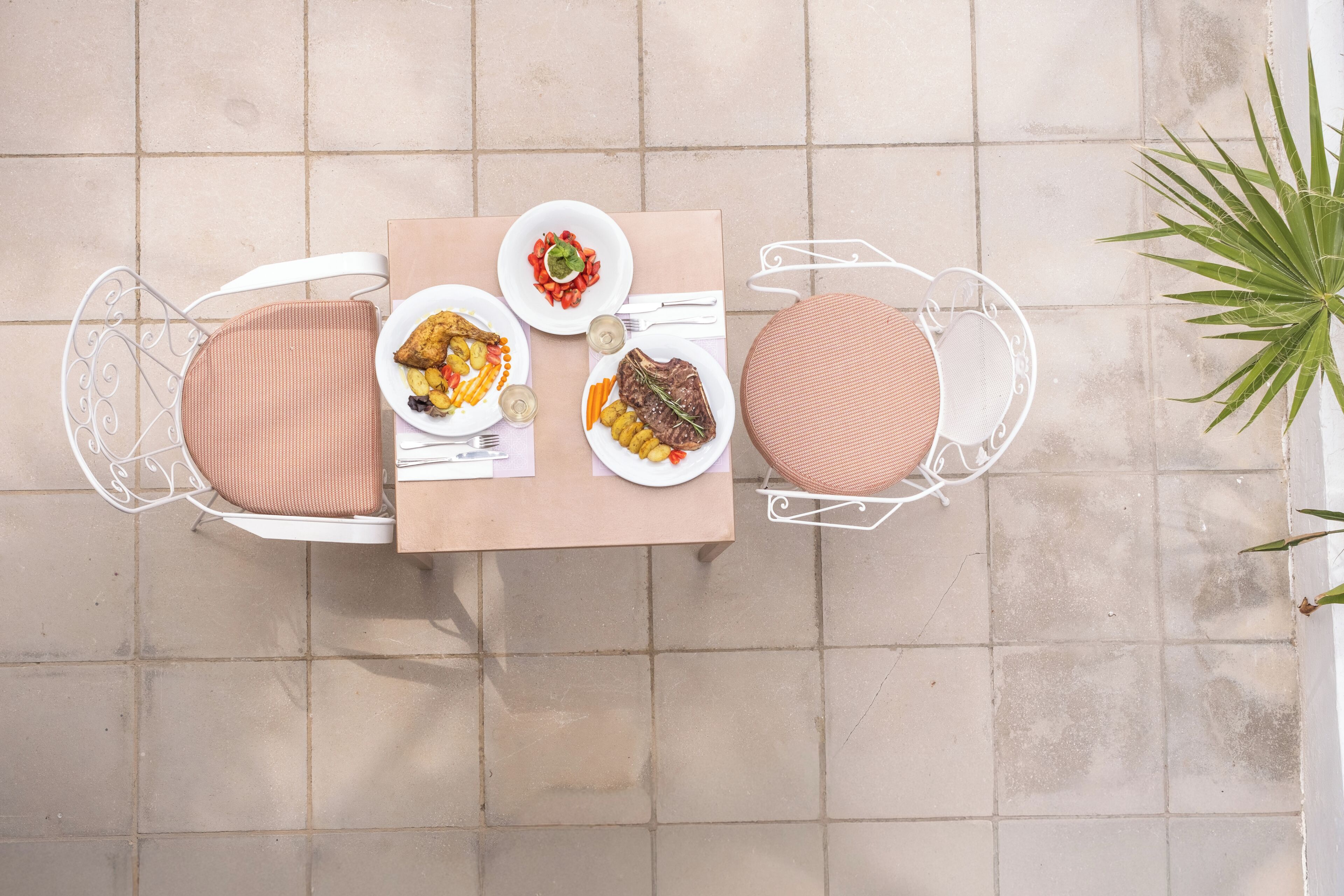A table with plates of food, white chairs, and a potted plant.