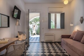 A room with a checkered floor, a sofa, a desk with a chair, a TV mounted on the wall, and a window with open shutters. - Rosebay Hotel (Santorini)
