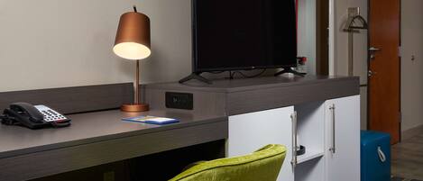 A hotel room with a desk, a television, a telephone, a lamp, a green chair, a wardrobe, and a blue cabinet.