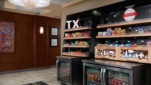 Un vestíbulo de hotel con un letrero 'TX', estantes de aperitivos y dispensadores de bebidas refrigeradas.