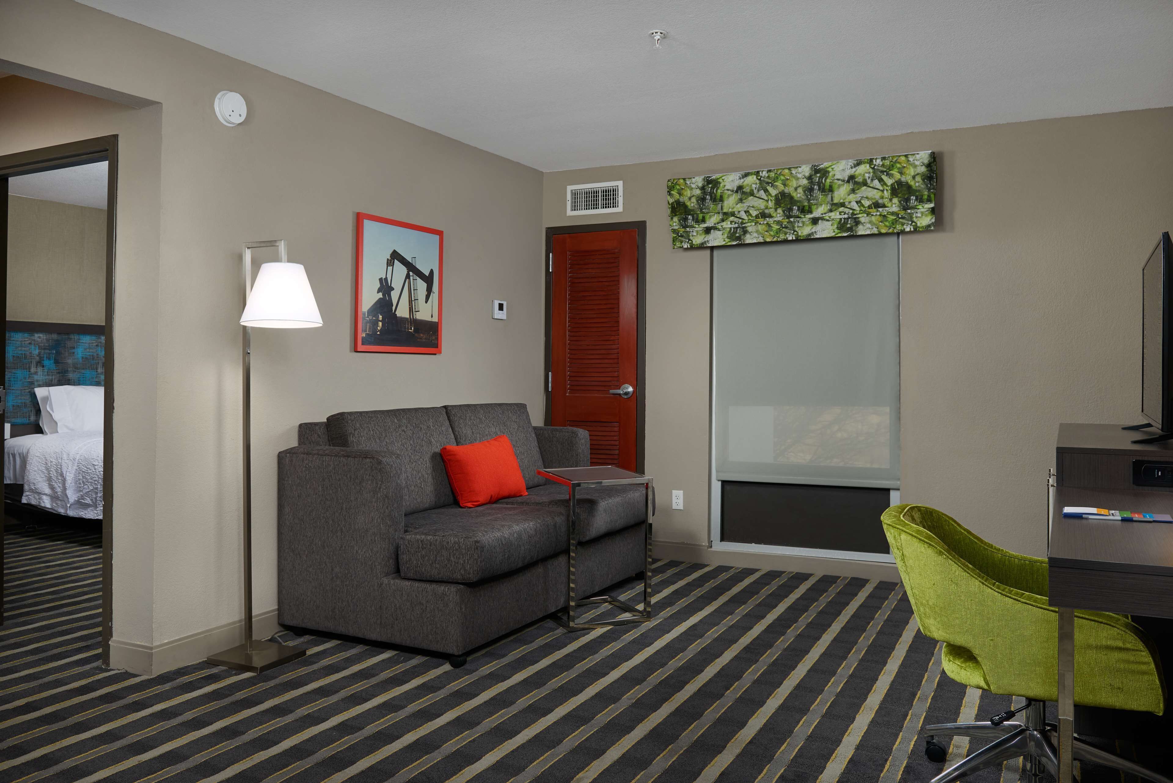 A hotel room with a gray sofa, a green chair, a desk, a lamp, a painting, and a door leading to another room.
