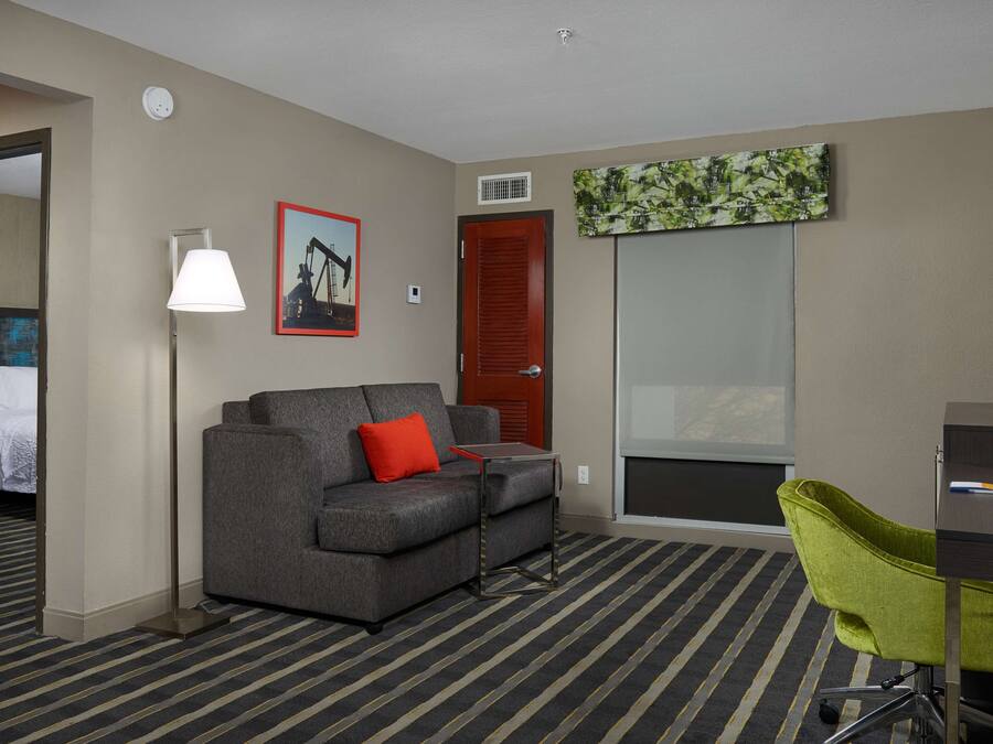 A hotel room with a gray sofa, a green chair, a desk, a lamp, a painting, and a door leading to another room.