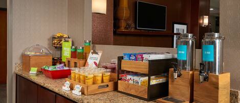 A hotel breakfast bar with juice dispensers, cereal boxes, and a fruit bowl.