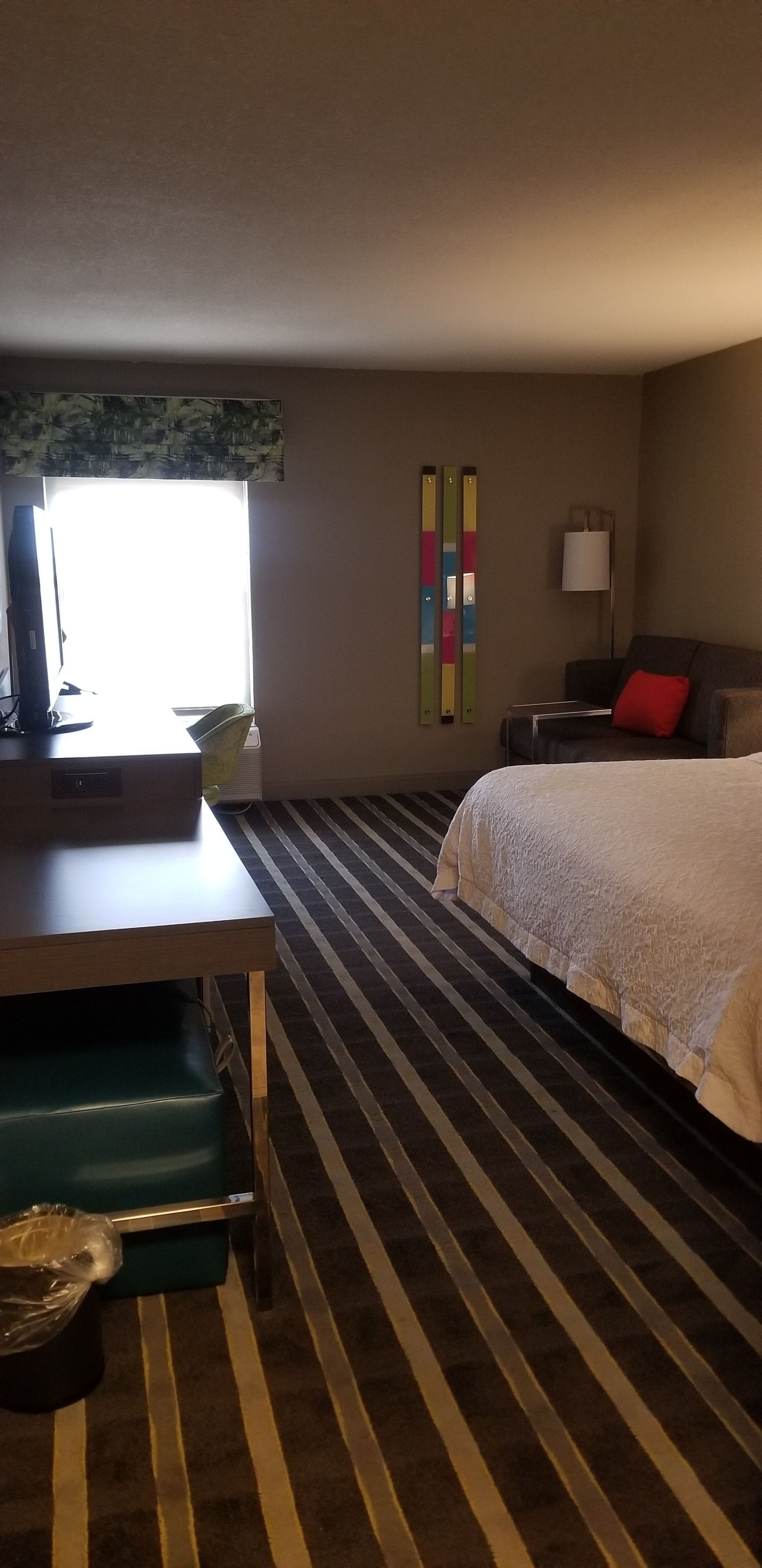 A hotel room with a striped carpet, a bed with a white cover, a desk with a computer, a chair, and a lamp.