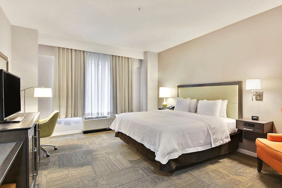 A hotel room with a large bed, a desk with a TV, a chair, and a window with curtains.